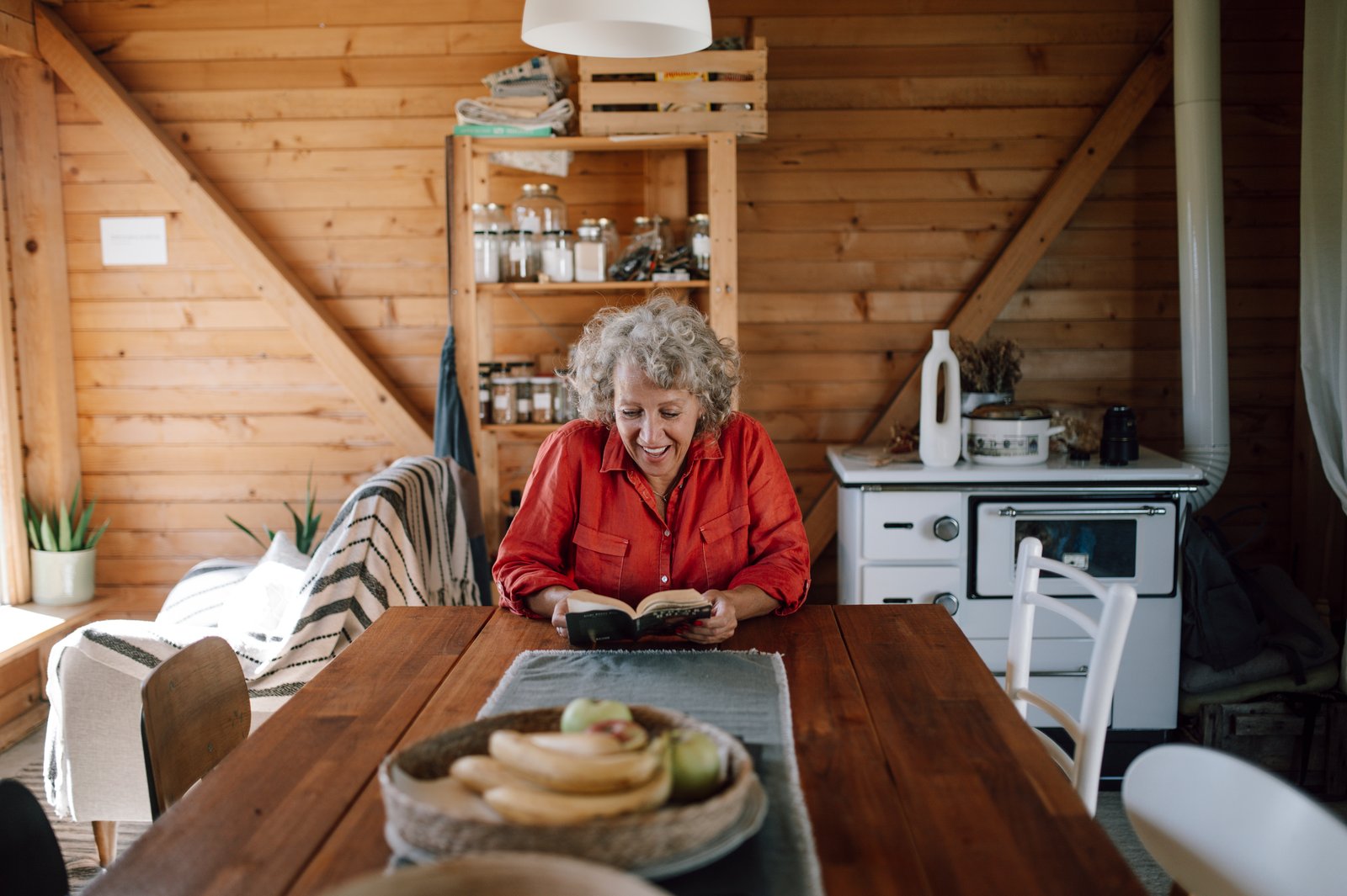 A senior woman smiles while reading a book at a wooden table in a cabin.